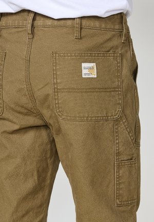 Hose aus olivgrünem Stoff mit einer aufgesetzten Tasche auf der Rückseite und einem Etikett mit gelben Akzenten in der Nähe des Bundes.