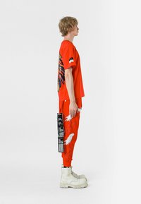 Camiseta roja con diseño de alas multicolores en la parte de atrás; joggers rojos con estampado de plumas blancas; botas de combate color crema; etiqueta negra adjunta.