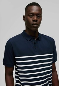 Marineblauwe polo met een witte horizontale streepjespatroon. Voorzien van een klassieke kraag, knoopsluiting en geborduurd merksymbool.