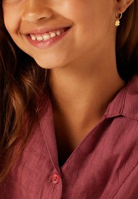 Chemise rose clair texturée avec un col et un bouton rose. Boucle d'oreille en or avec un charme en forme de trèfle. Peau lisse et cheveux marron ondulés.
