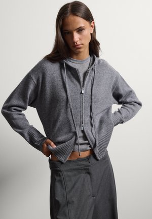 Giovane donna con lunghi capelli castani che indossa una felpa grigia con zip sopra un maglione a coste e pantaloni larghi grigio scuro su uno sfondo neutro.