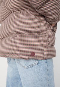 Casaco puffer em padrão xadrez castanho e rosa com um acabamento texturizado. Apresenta um logótipo redondo em bordô na borda inferior.