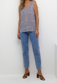 Ärmelloses Oberteil mit V-Ausschnitt aus hellblauem Stoff, der ein braunes Streifenmuster aufweist. Kombiniert mit hellblauen Jeans und sandfarbenen Blockabsatzsandalen.