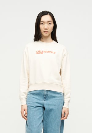 Žena nosi kremasti pulover s natpisom "KARL LAGERFELD JEANS" narančastim slovima i svijetloplave traperice, stojeći uz jednobojnu svijetlu pozadinu.