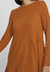 Robe en maille côtelée couleur rouille avec manches longues et détail taille basse portée par une personne sur un fond uni.