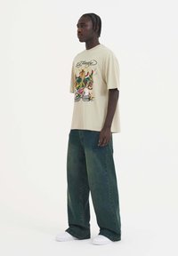Camiseta gráfica beige con un estampado colorido, combinada con pantalones de pierna ancha verde oscuro y zapatillas blancas. Diseño sencillo, corte relajado.