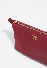 Funda de tela roja con cremallera dorada, con el logotipo en relieve "BIMBA Y LOLA". Textura suave, forma rectangular y vista lateral en ángulo.