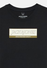 Camiseta negra con un logotipo rectangular que dice "JACK & JONES" y "DESDE MIL NOVECIENTOS NOVENTA" en tonos beige y verde olivo.