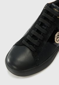 Baskets noires avec un embout en cuir lisse, tige en daim, lacets noirs et un détail en métal sur le côté. Surface texturée.