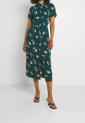 Femme portant une robe midi verte à fleurs boutonnée, manches courtes et ceinture à la taille, associée à des sandales noires à talons.