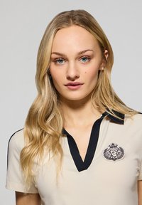 Camisa polo beige con un cuello negro y un diseño de escote en V. Presenta un logo bordado en el lado izquierdo del pecho y mangas cortas.