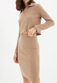 Žena oblečená v žebrovaném béžovém dvoudílném outfitu s krátkým rukávem a midi sukní s vysokým pasem na bílém pozadí.