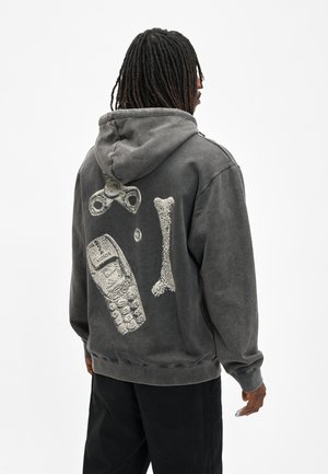 Man draagt een vervaagde zwarte hoodie met witte afbeelding van een spinner, bot en oude mobiele telefoon op de rug, staand met zijn rug naar voren.