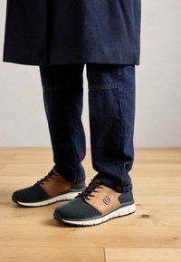 Personne portant un pantalon en denim bleu et des baskets décontractées marron et noires, debout sur un sol en bois.