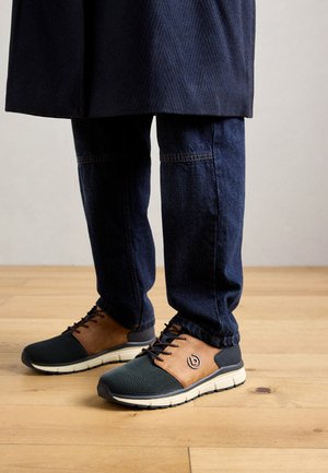 Egy kék farmernadrágot és barna-fekete laza sportcipőt viselő személy áll egy fából készült padlón.