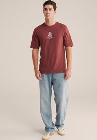 Mørkerød kortærmet t-shirt med hvid grafisk print på brystet, parret med lyseblå løst sidende denim jeans og hvide sneakers.