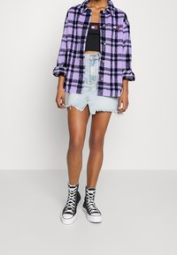 Fialová a černá kostkovaná košile-jacket, černý crop top, roztrhaná džínová sukně a černé vysoké tenisky. Viditelné detaily knoflíků a třepení.