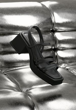 Black heeled sandal with multiple thin straps, square toe, and a chunky heel. Smooth texture on a metallic surface background.