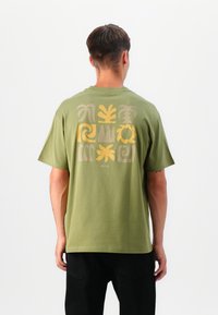 Olīvu zaļš kokvilnas t-krekls ar daudzkrāsainu ģeometrisku rakstu aizmugurē, ar īsām piedurknēm un brīvu piegriezumu.