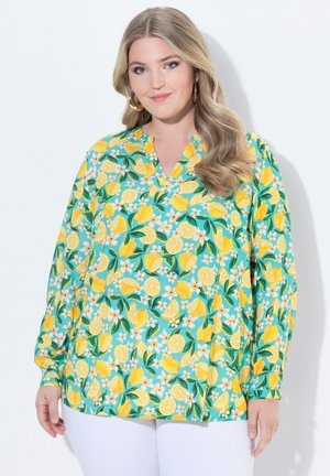 Frau trägt eine langärmelige blaugrüne Bluse mit gelbem Zitronen- und weißem Blumenmuster, kombiniert mit weißen Hosen, steht vor weißem Hintergrund.