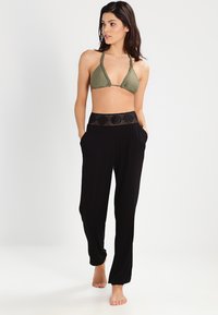 Top de bikini triangular en verde oliva con tirantes trenzados, combinado con pantalones negros holgados que tienen una cintura texturizada con patrón y bolsillos laterales.