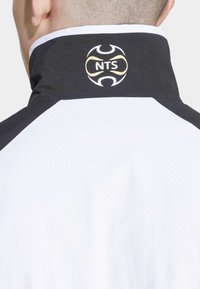 Vēja mētelis ar melnu apkakli un zelta "NTS" logotipu, balts korpuss ar tekstūrētu audumu un kontrastējošiem melniem akcentiem plecos.
