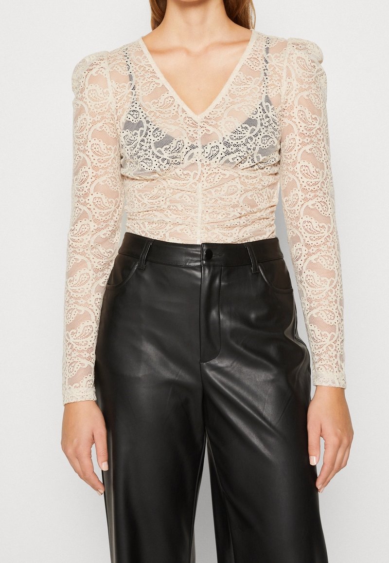 Femme portant un chemisier beige en dentelle à manches longues sur un soutien-gorge noir, associé à un pantalon en cuir noir taille haute sur un fond uni.