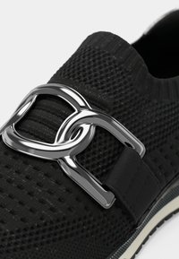 Μαύρο αθλητικό παπούτσι φτιαγμένο από υλικό πλεκτού με υφή, με έντονο ασημί μεταλλικό στοιχείο αλυσίδας και μαύρο ελαστικό λουρί για καλή εφαρμογή.