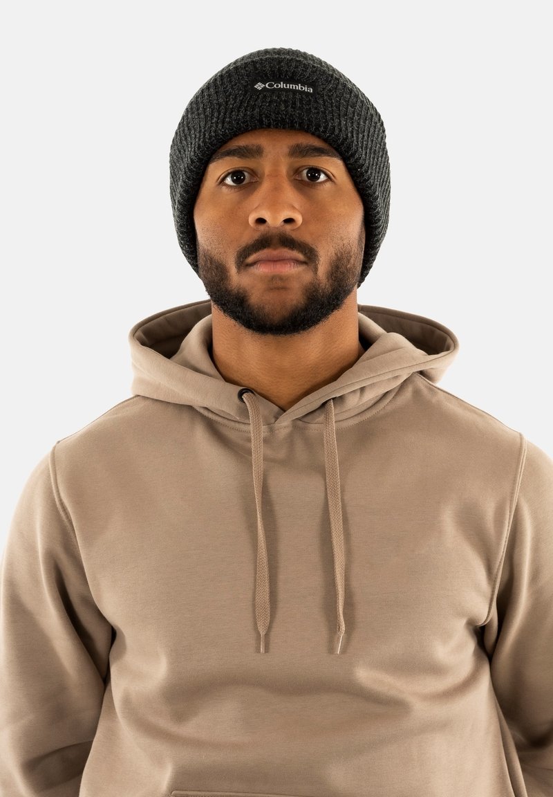 Gorro de punto gris oscuro con un logo gris claro, llevado con una sudadera beige que presenta un bolsillo canguro y detalles de cordón.