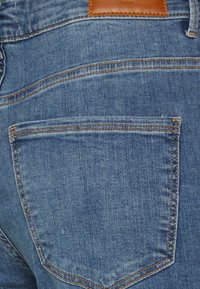 Jeans de ganga azuis com um bolso traseiro com costura contrastante e uma etiqueta de couro castanho acima dele. O tecido tem uma aparência lisa e ligeiramente texturizada.