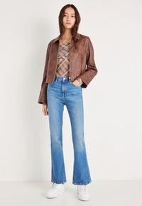 Chaqueta corta de cuero marrón con cierre de cremallera, combinada con vaqueros acampanados azul claro y zapatillas blancas. La chaqueta tiene un diseño con cuello.