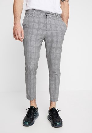Man i grå rutiga slim-fit byxor, en vit skjorta instoppad, svarta sneakers, står med en hand i fickan.
