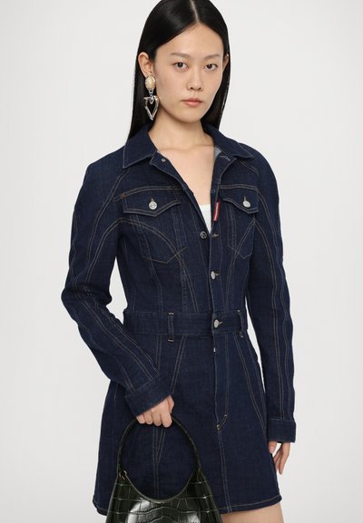 Dark blue denim dress with long sleeves, snap buttons, front pockets, and a belted waist. Model holds a black handbag.