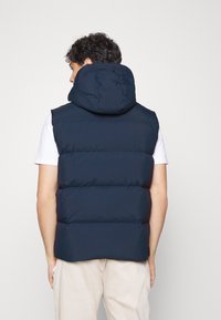 Mörkblå vadderad väst med huva, med ett quiltat mönster och synliga sömmar. Baktill syns en hög krage och ärmlös design.