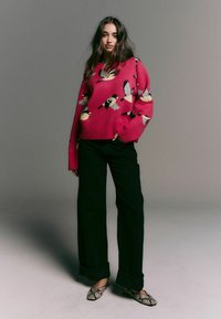 Rosa Strickpullover mit Vogelmustern, weiten Ärmeln und einem gerippten Saum, kombiniert mit schwarzen weit geschnittenen Hosen und gemusterten Schuhen.