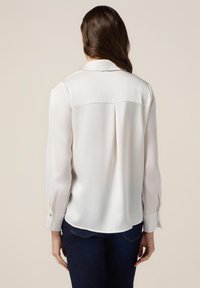 Blusa di seta bianca con maniche lunghe, colletto, polsini con bottoni e una piega centrale sul retro. Vestibilità slim, orlo arrotondato, abbinata a jeans scuri.