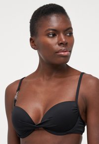 Černý bikiny top s krouceným designem, hladkou texturou, nastavitelnými ramínky a dekoračním kovovým prvkem na levém ramínku.