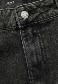 Detail čierneho denimového materiálu, ktorý zobrazuje viditeľnú textúru s diagonálnymi pruhmi, strieborný gombík s nápisom "NEXT DENIM" a kontrastné stehy.