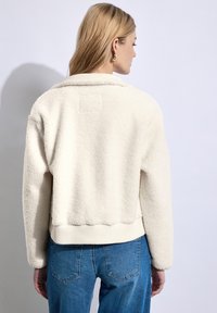 Jonge vrouw met blond haar die een crèmekleurige fleece jas en een blauwe spijkerbroek draagt, met haar rug naar de camera toegekeerd tegen een effen achtergrond.