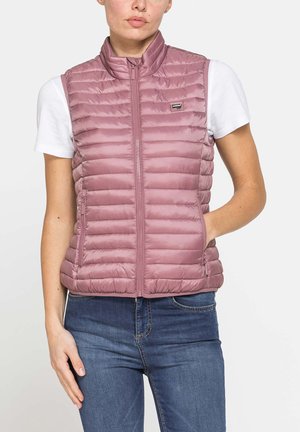 Donna che indossa un gilet trapuntato mauve senza maniche sopra una camicia bianca a maniche corte e jeans blu, con una mano nella tasca del gilet.