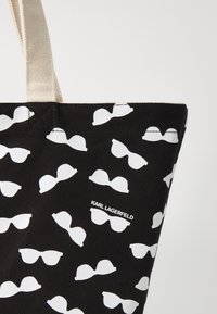 Juodas tote krepšys su pasikartojančiu baltų saulės akinių raštu. Pagamintas iš kanvos medžiagos su smėlinėmis rankenomis. Logo "KARL LAGERFELD" atspausdintas baltai.