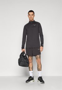 Svart langermet treningstopp med glidelås, kombinert med svarte shorts. Modellen holder en svart treningsbag og har på seg svarte sko med grønne detaljer.