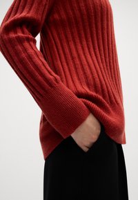 Czerwony sweter prążkowany z wydłużonymi rękawami, posiadający teksturowany wzór i luźny krój. Częściowo widoczna czarna odzież pod nim.