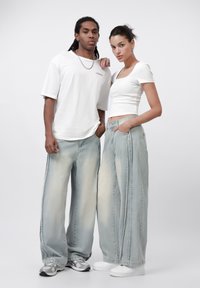 Två modeller bär stora ljusblå denimjeans med blekta områden. Den manliga modellen har en lös vit T-shirt, och den kvinnliga bär en figurnära vit topp.