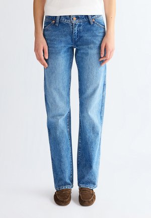 Osoba u ravnim plavim trapericama, bijeloj košulji i smeđim cipelama u stilu mokasina, stoji na svijetloj pozadini.