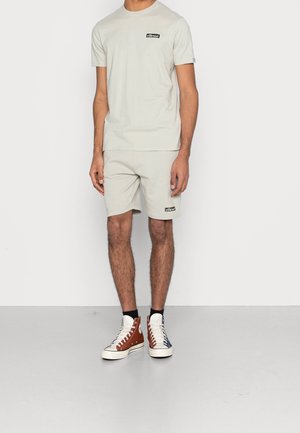Uomo che indossa una t-shirt beige chiaro a maniche corte e pantaloncini abbinati con loghi neri, abbinati a sneakers alte bicolore e calzini neri.