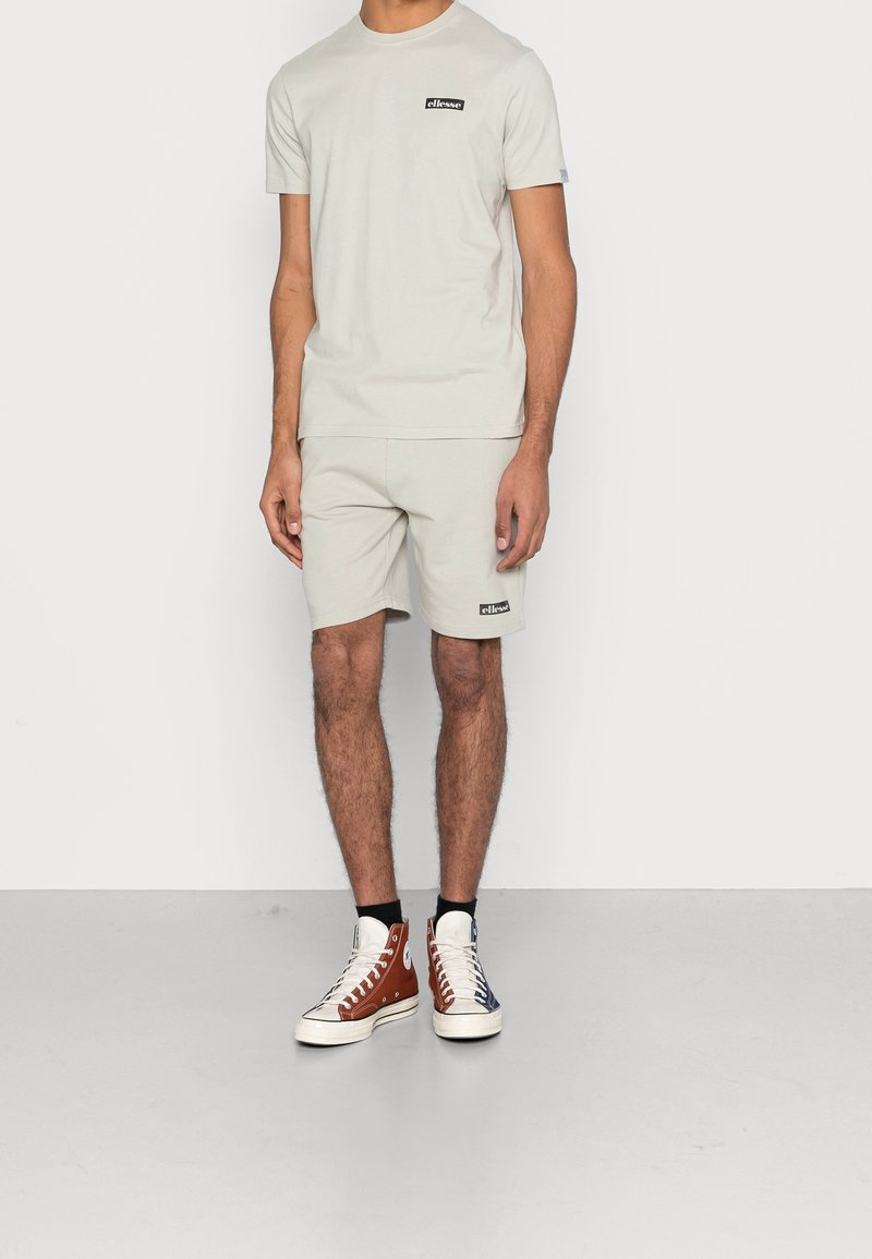 Homme portant un t-shirt à manches courtes beige clair et un short assorti avec des logos noirs, accompagné de baskets montantes bicolores et de chaussettes noires.