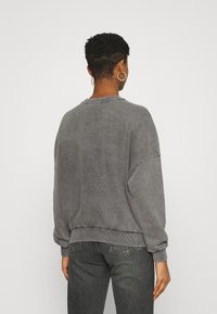 Persona con capelli corti che indossa un grande sweatshirt grigio e jeans scuri, in piedi contro uno sfondo chiaro e semplice, girata di spalle.