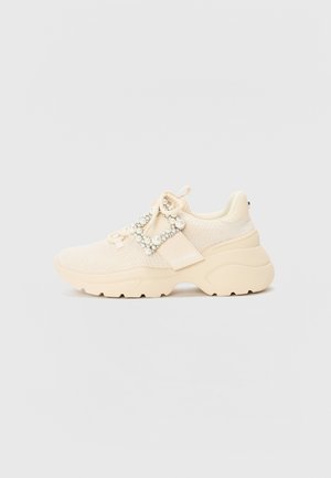Beige Sneaker mit einem strukturierten Strick-Obermaterial, dicker Gummisohle und dekorativen Perlenakzenten auf den Schnürsenkeln für zusätzliche Details.