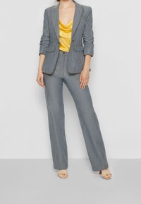 Completo in denim grigio con blazer su misura dotato di rever a punta e due tasche frontali, abbinato a pantaloni a gamba larga e una blusa in satin gialla.